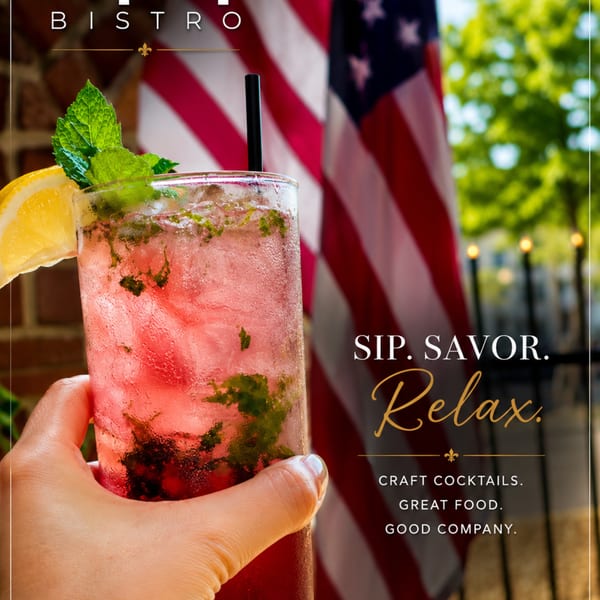 Hand holding a red cocktail with mint and lemon, an American flag and green trees in the background.