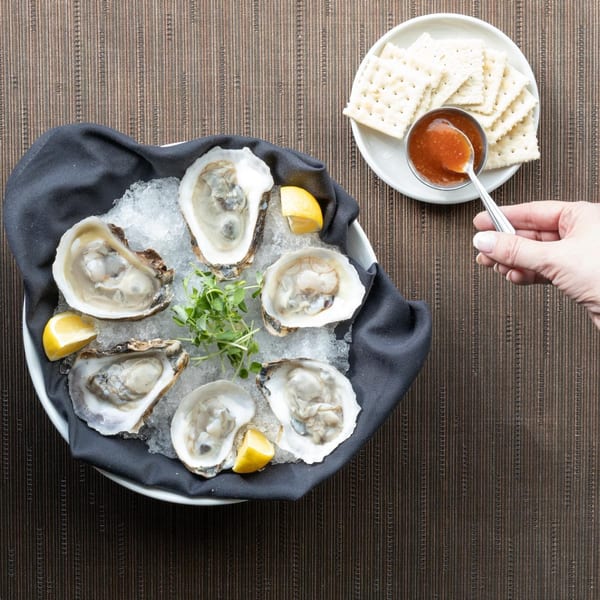 Six fresh oysters on ice with lemon wedges, microgreens, and a side of cocktail sauce and crackers.