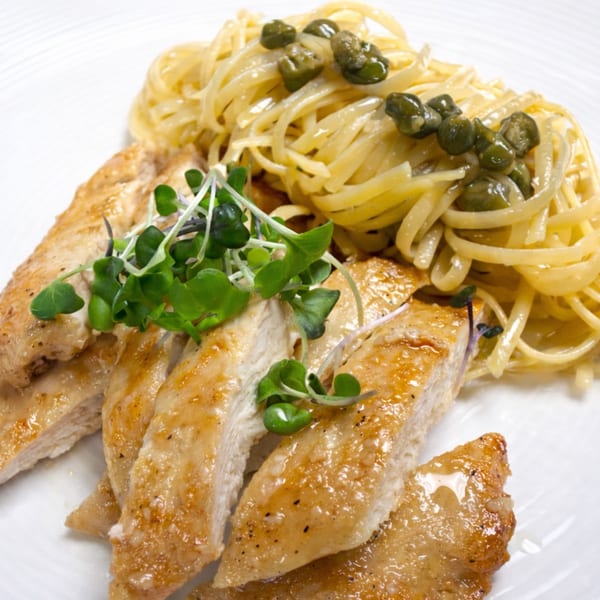 Chicken piccata with caper-topped linguine and microgreens on a white plate.