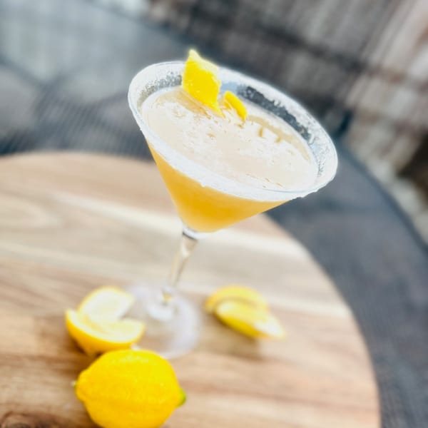 Lemon drop martini with a sugared rim and lemon peel garnish, with fresh lemons on a wooden table.