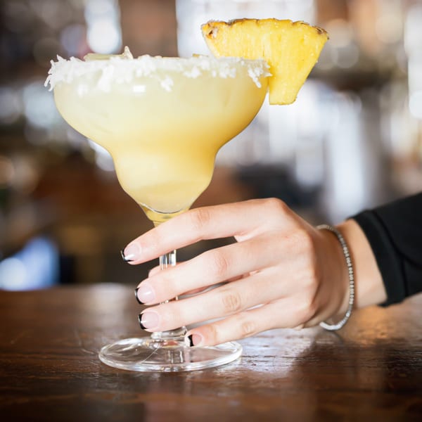 A hand with black-tipped nails holds a pineapple margarita with a coconut-flaked rim.