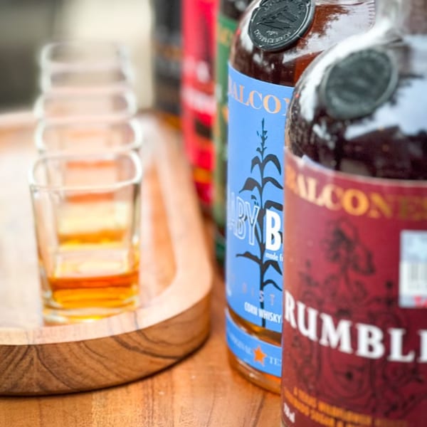 Four shot glasses filled with amber liquid on a wooden tray next to two bottles of spirits.