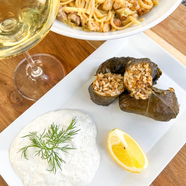 Dolmades with tzatziki, lemon, and a glass of white wine, with pasta in the background.