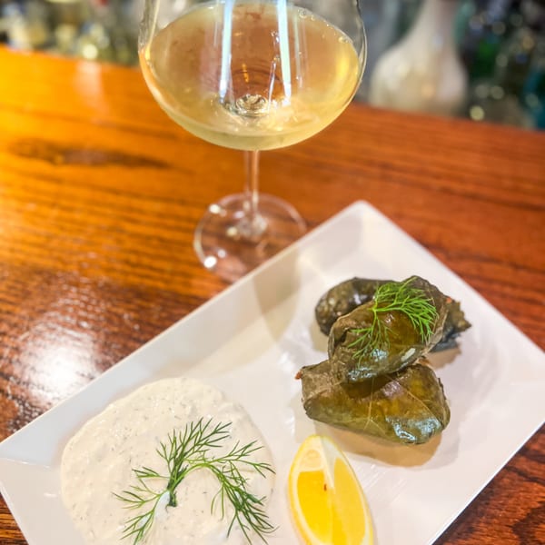 Grape leaves, tzatziki, and a lemon wedge on a white plate with a glass of white wine.