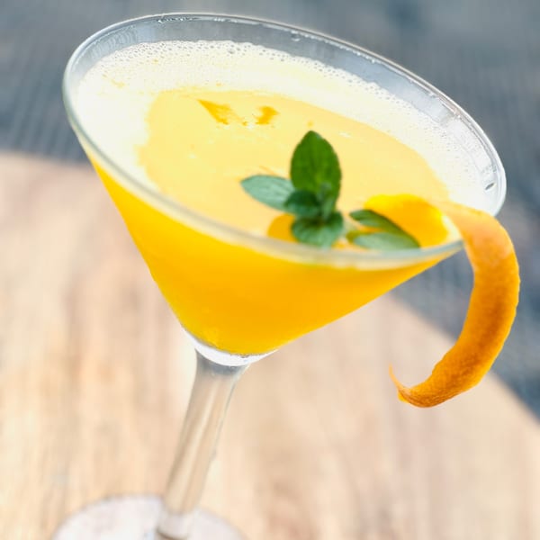 A vibrant yellow cocktail in a martini glass, garnished with mint leaves and an orange peel.