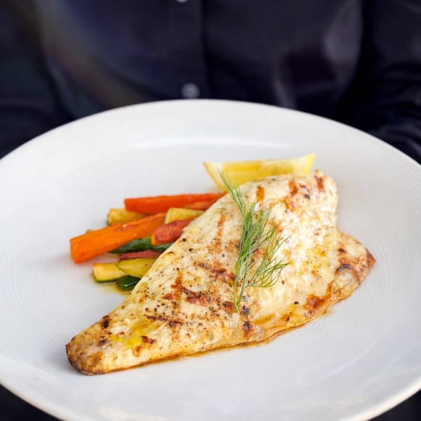 Grilled fish fillet with roasted carrots, zucchini, a lemon wedge, and dill on a white plate.