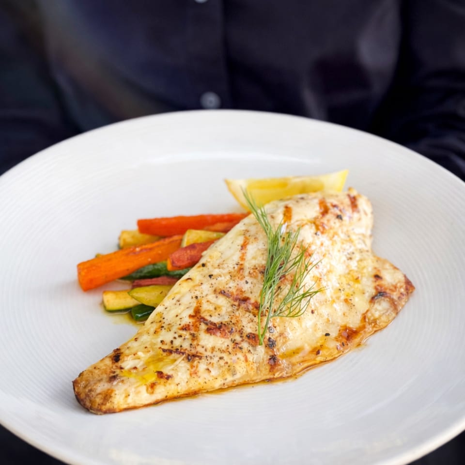 Grilled fish fillet with roasted carrots, zucchini, a lemon wedge, and dill on a white plate.