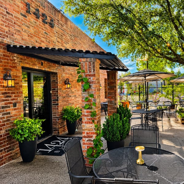 Brick building with a black awning, outdoor patio seating, and lush green plants under a sunny sky.